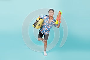 Happy handsome Asian tourist man in summer outfits with water gun and suitcase in blue color studio isolated background