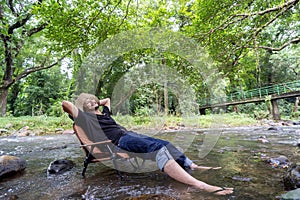 Happy handsome bearded man wearing a hiking hat resting on a camping chair in a stream in the forest and enjoying the surrounding
