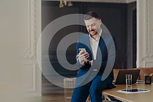 Happy handsome businessman looking at mobile phone while sitting on office desk