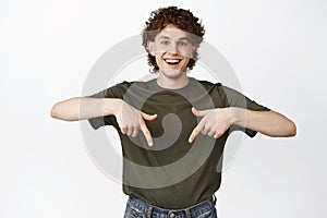 Happy handsome man pointing fingers down and laughing, showing advertisement, standing in tshirt over white background