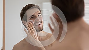 Happy handsome metrosexual guy applying sunscreen on face at mirror