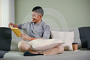 Happy and handsome millennial Asian man reading book or novel on sofa in his living room
