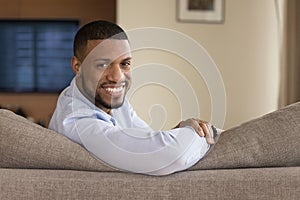 Happy handsome young Black homeowner man sitting on couch