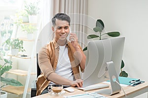 Happy handsome young businessman using computer and talking on cell phone in office
