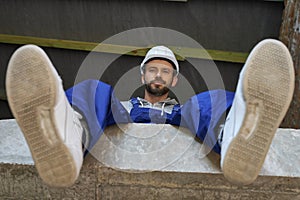 Happy handsome young male builder in blue overalls and hard hat looking at camera, sitting on the concrete floor while