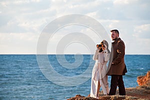 Happy handsome young pair trendy bearded man and woman in autumn coat looking through binoculars near sea shore during sunset with