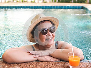 Happy and healthy Asian senior  woman wearing straw hat and sunglasses  drinking  orange juice  in the swimming pool, smiling and