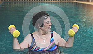 Happy and healthy senior Asian woman  working out in the swimming pool with yellow dumbbells, smiling and looking away. Elderly