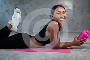 Happy healthy smiling woman holding protein shake relaxing on the pink yoga mat at the gym