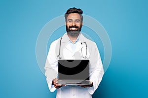 Happy hindu man doctor in white coat with stethoscope showing laptop with blank screen on blue studio background