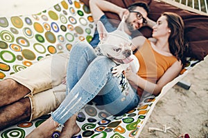 Happy hipster couple with bulldog relaxing in hammock on the beach in sunset light, summer vacation. stylish family with dog