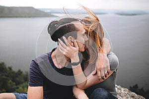 Happy hipster couple kissing on top of rock mountain with beautiful view on river. Tourist couple sitting and hugging on windy