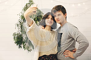 Happy hipster couple taking selfie at modern christmas tree lights in stylish festive room. Celebrating Christmas or New Year`s
