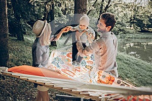 Happy hipster family playing with cute little son on hammock in summer sunny park. stylish mom and dad with kid relaxing in forest