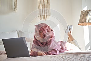 Happy hipster gen z teen girl with pink hair watching movie on laptop in bed.