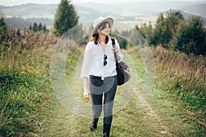 Happy hipster girl with backpack traveling on top of sunny mountain, walking on hills with woods. Stylish woman smiling and