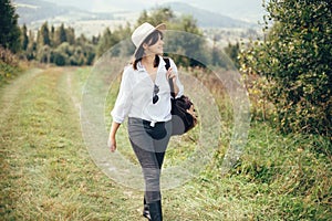 Happy hipster girl with backpack traveling on top of sunny mountain, walking on hills with woods. Stylish woman smiling and