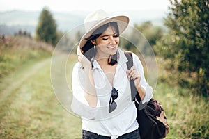 Happy hipster girl with backpack traveling on top of sunny mountain, walking on hills with woods. Stylish woman smiling and