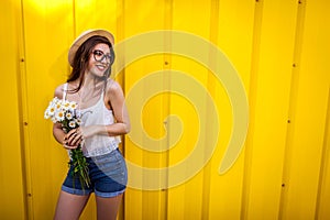 Happy hipster girl wearing glasses and hat with flowers against yellow background. Summer outfit. Copy Space.