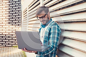 Happy hipster guy using his laptop while standing outdoors on a sunny day