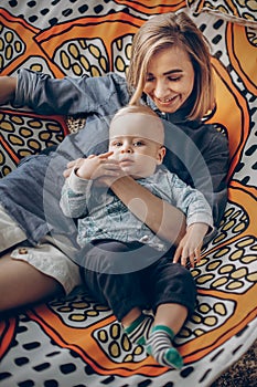 Happy hipster mother playing with her cute little son on hammock in summer sunny park. stylish mom with kid relaxing in forest and