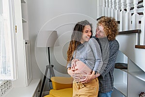 Happy hipster pregnant couple standing together near stairs in contemporary apartment and tenderly touching belly.