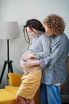 Happy hipster pregnant couple standing together near window in contemporary apartment and tenderly touching belly.