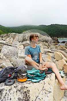 Happy hipster tourist young woman enjoying halt sitting on stone beach with portable camp gas stove