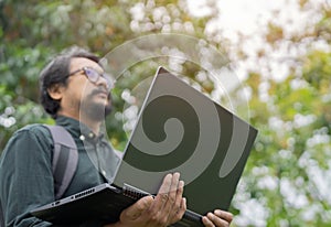 Happy hipster young Asian man wearing glasses standing and working on laptop in the park