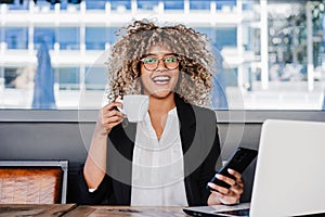 Happy hispanic afro business woman wearing eye glasses in cafe working on laptop, mobile phone