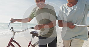 Happy hispanic senior couple walking with bikes on beach at sunset