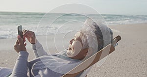 Happy hispanic senior woman relaxing on sunbed on beach at sunset, using smartphone