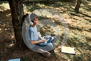 Happy Hispanic teenage boy in headphones, relaxing in the park after school, sitting under tree near scattered textbooks