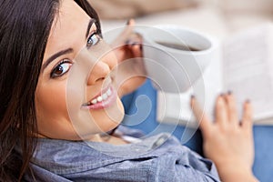 Happy Hispanic Woman Reading Book at Home and Drinking Tea or Coffee