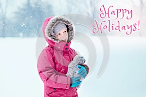 Happy holidays. Christmas card with text. Caucasian smiling girl child looking at  falling snow on cold winter snowy day outdoors