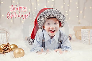 Happy holidays. Christmas card with text. Cute adorable Caucasian boy in Santa hat lying on a white fluffy carpet on ground.