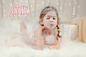 Happy holidays. Christmas card with text. Cute adorable Caucasian girl in princess diadem crown lying on white fluffy carpet on a