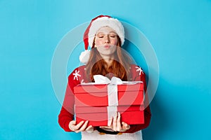 Happy holidays and Christmas concept. Cute redhead girl holding presents and pucker lips for kiss, wearing santa hat and