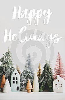 Happy holidays Greeting card. Happy holidays text handwritten on christmas little houses and trees on white background,  winter