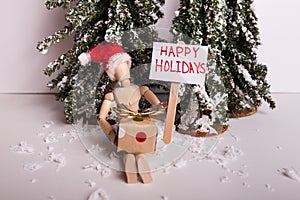 Happy Holidays picket sign held by wooden jointed manikin doll with wrapped gift wearing a Santa Claus hat wintery scene