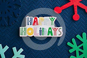 Happy Holidays plaque message and colorful wooden snowflakes on blue background, for Christmas projects