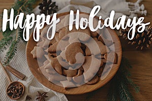 Happy Holidays text on christmas gingerbread cookies in wooden plate with fir branches and cones on table. Merry Christmas.