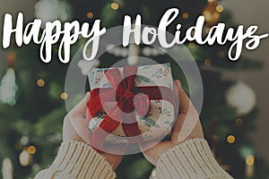 Happy Holidays text on hands in cozy sweater holding stylish christmas gift with red ribbon at christmas tree with lights. Merry