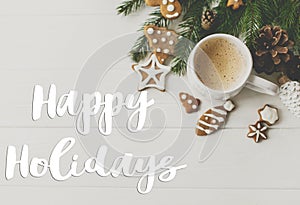Happy holidays text handwritten on christmas flat lay with gingerbread cookies, coffee, pine cones and fir branches on white wood