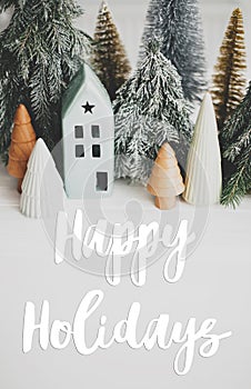 Happy holidays text handwritten on christmas little houses and trees on white background,  winter holiday village scene. Greeting