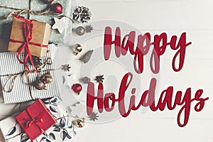 happy holidays text, seasonal greetings card sign. flat lay. presents with ornaments pine cones anise on rustic white wooden