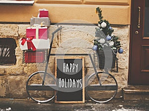 Happy Holidays text sign on board and stylish bicycle with christmas gifts and modern christmas tree at store front at holiday