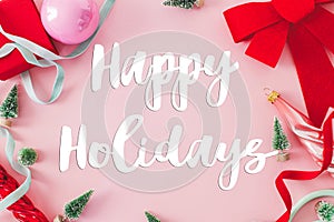 Happy Holidays text sign on christmas red bow, baubles, little green trees and ribbons on pink background flat lay. Season`s