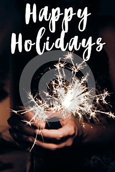 happy holidays text sign, greeting card. happy new year and merry christmas concept. female hand holding a burning sparkler