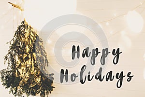 Happy Holidays text sign on modern christmas tree made of pine branches with golden festive lights hanging on white wooden wall.